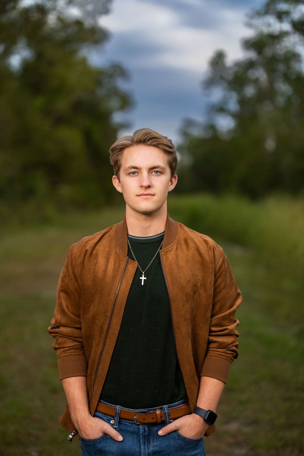 Person in brown jacket and jeans stands casually in outdoor setting with hands in pockets.