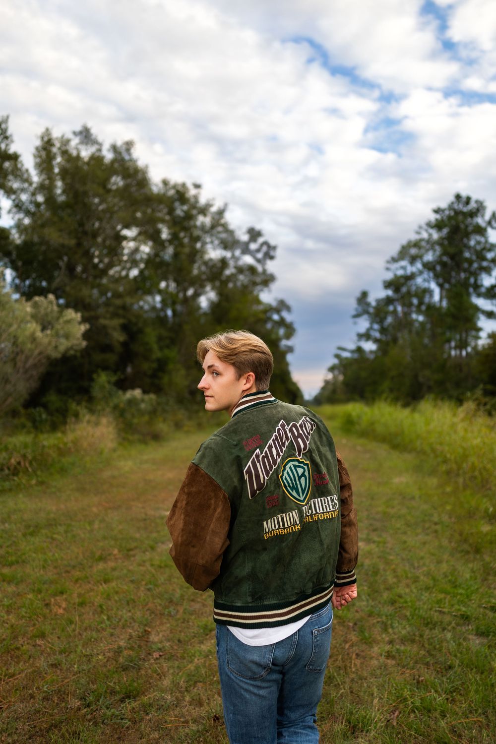 Person in varsity jacket shown from behind on grassy hill under cloudy sky.