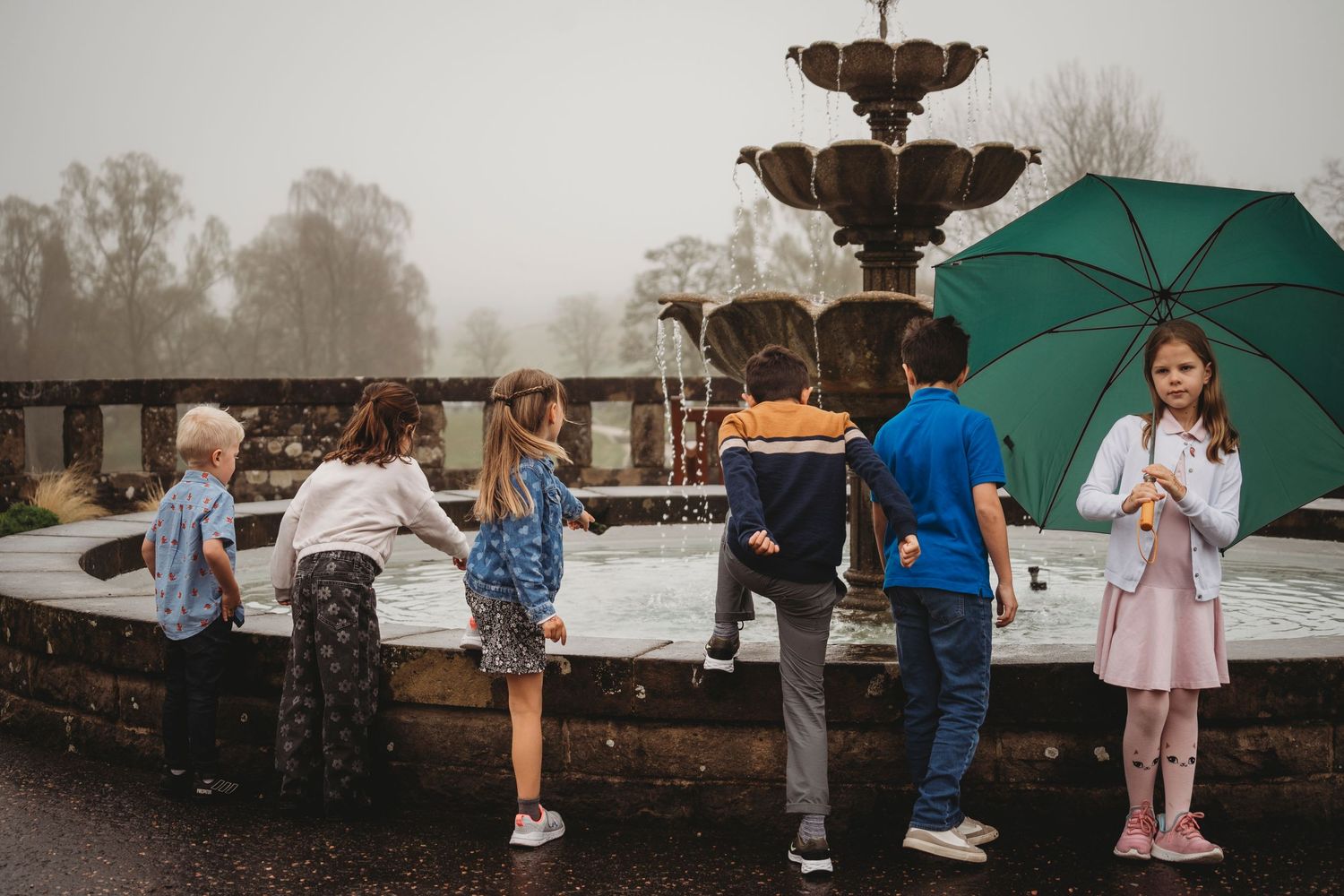 Group of people standing at a stone railing on a rainy day with a green umbrella and fountain in the background.