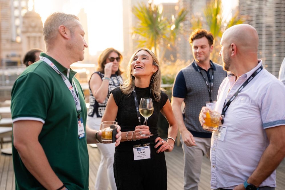 Professionals networking at rooftop event, woman in black dress laughing with wine glass, golden hour backdrop.