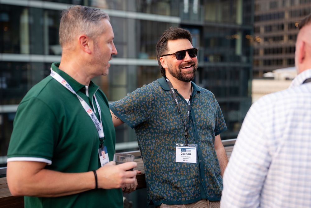Three men wearing conference lanyards smile and chat outdoors at a professional networking event.