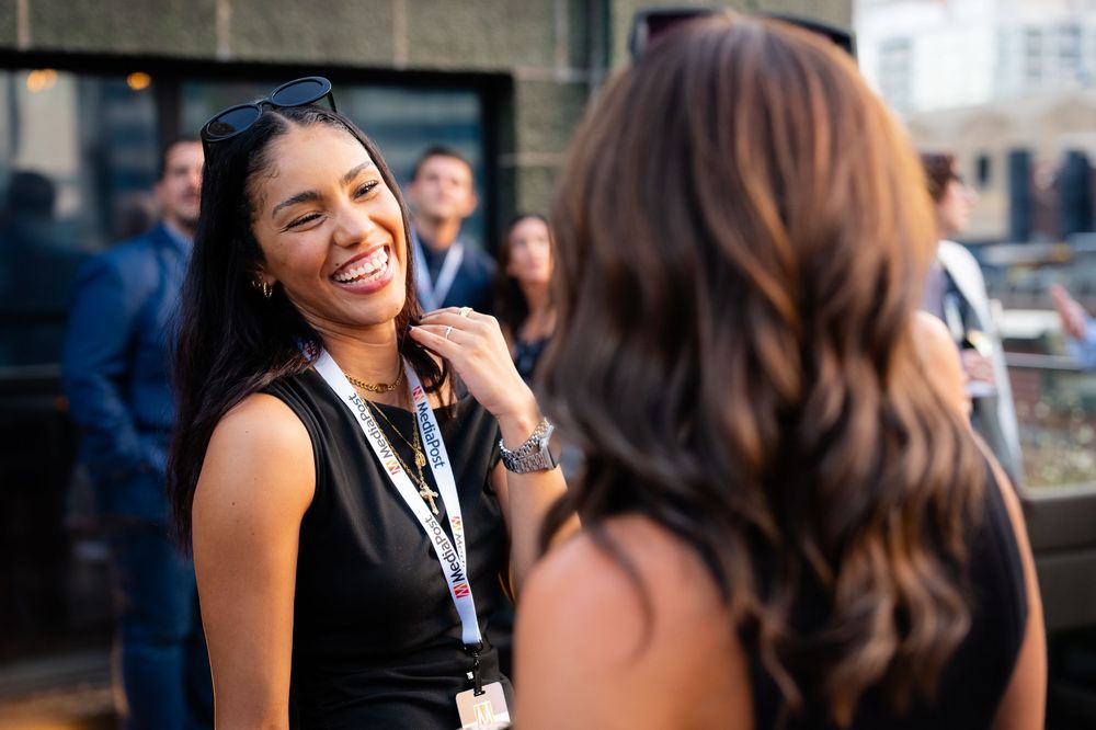 Two women in black outfits smile and chat at an outdoor networking event with city skyline in background.