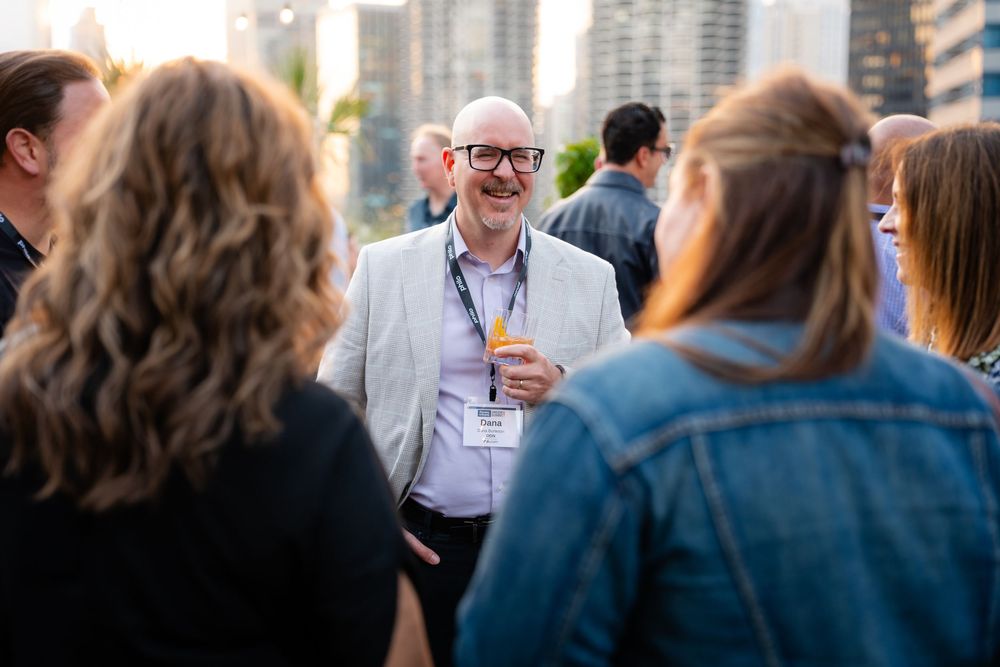 Man in gray blazer laughs while networking with colleagues at an outdoor rooftop business event.