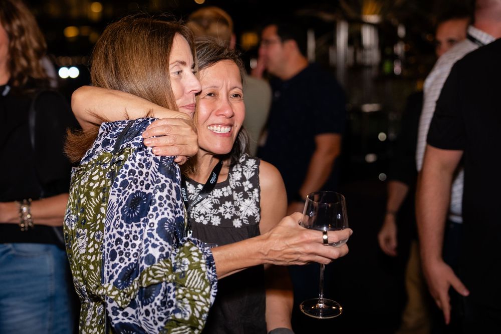Two women sharing a warm hug at a social gathering, one holding a glass of red wine, in a dimly lit venue.