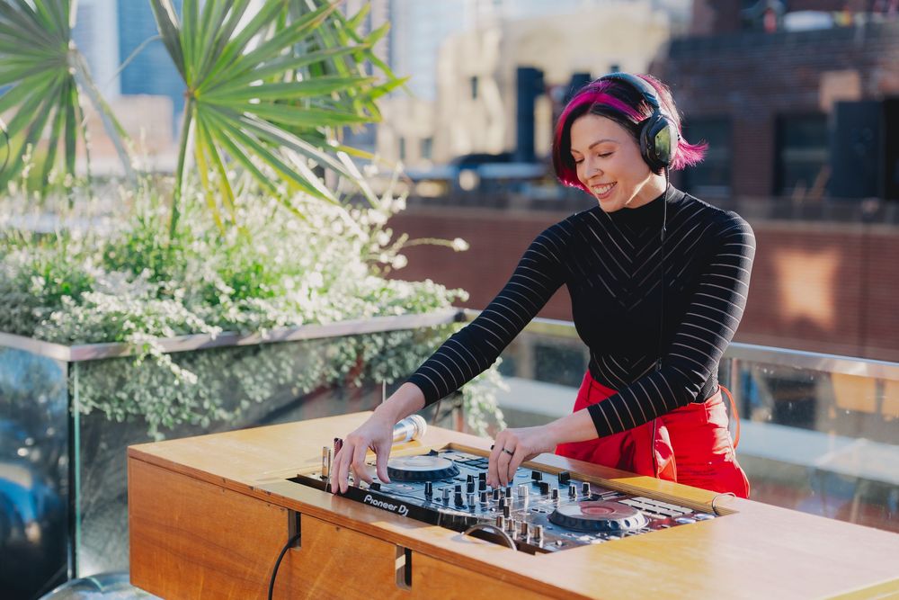 A female DJ with pink hair and headphones mixing music on a rooftop deck on a sunny day.