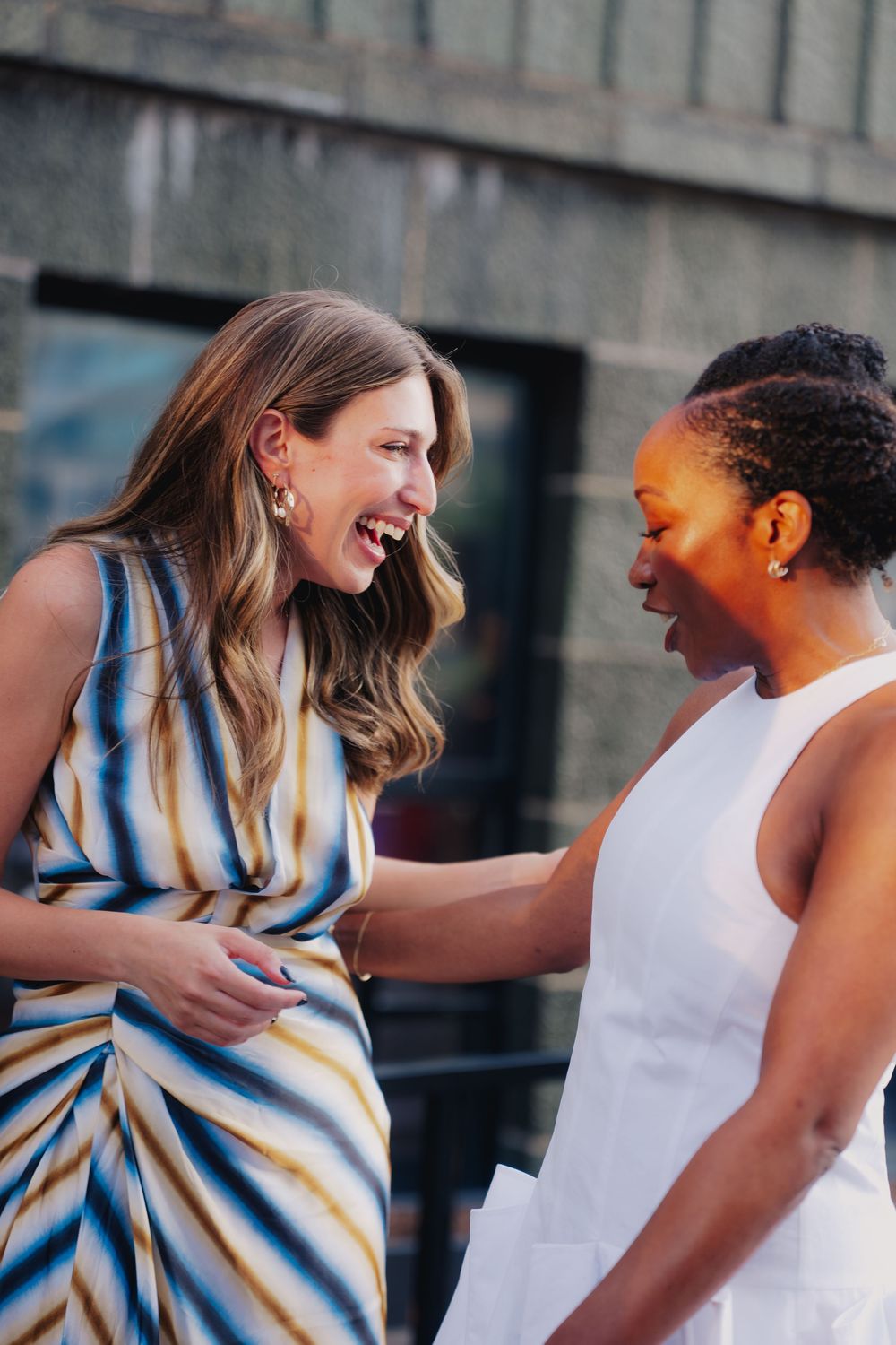 Two women laughing and sharing a joyful moment outdoors, one in a striped dress and one in white.