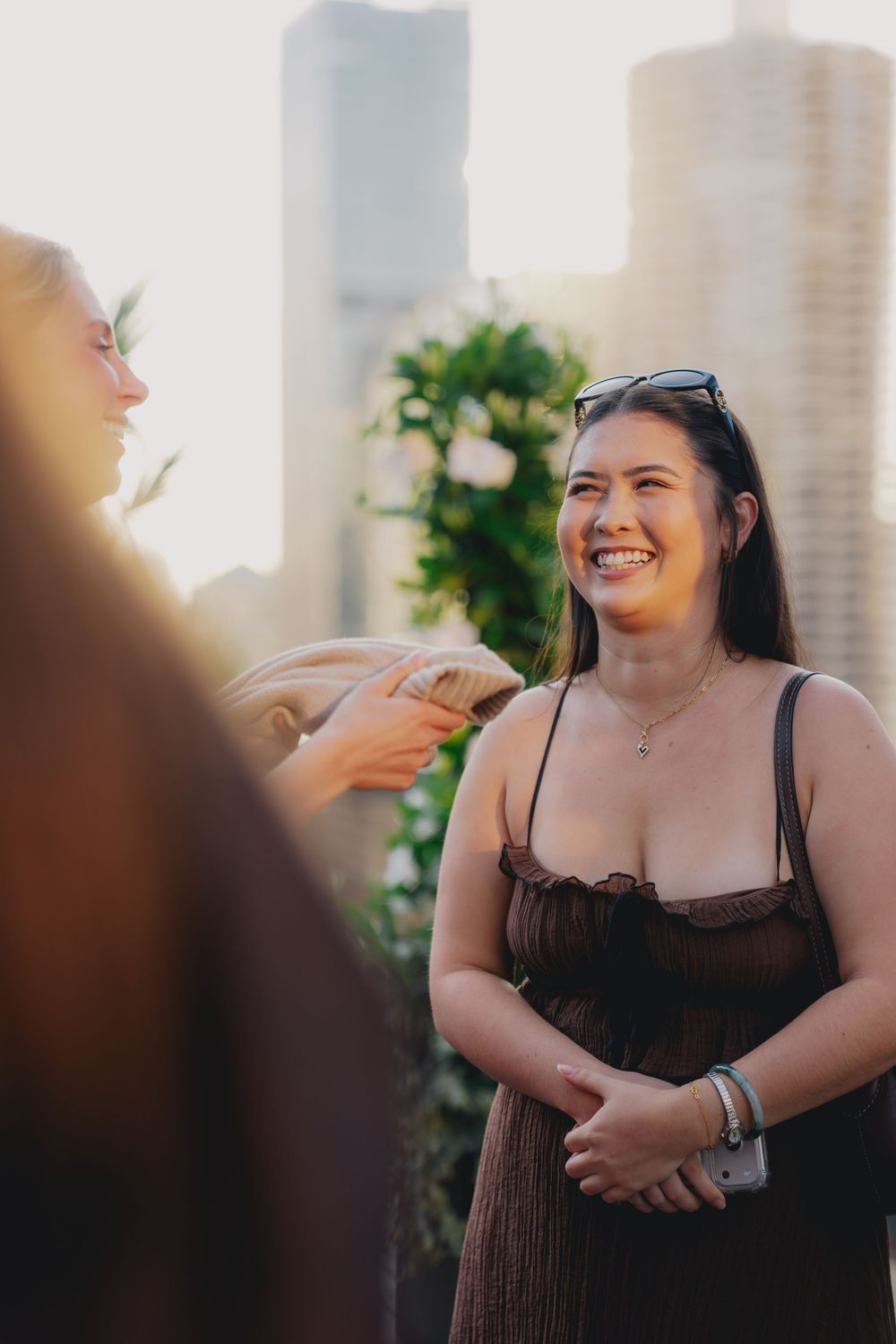 Smiling young woman in a brown dress with sunglasses on her head, chatting outdoors with city skyline in background.