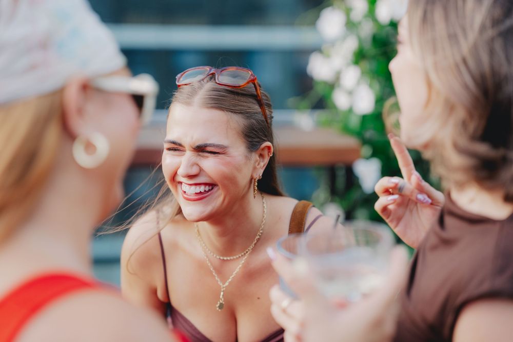 Three women laughing and socializing outdoors, holding cocktails at a sunny rooftop gathering.