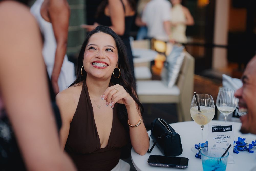 A smiling woman in a brown halter dress sits at a table with wine glasses at an elegant outdoor event.