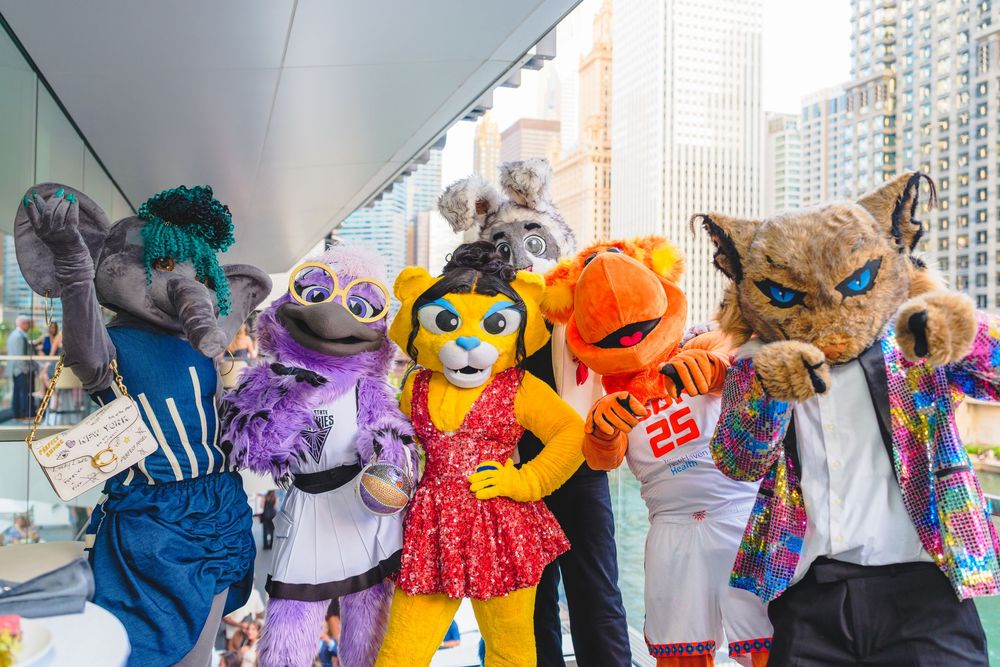 Six colorful animal mascot costumes pose together outdoors in a city setting on a sunny day.
