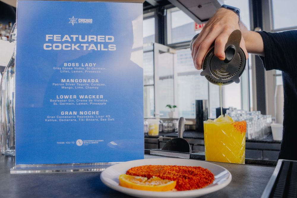 Featured cocktails menu display with orange drink and snack plate at a bar, bartender pouring a beverage.