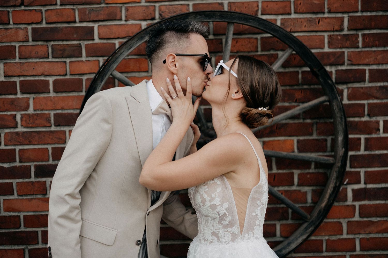 Couple shares romantic kiss against rustic brick wall with vintage wheel decor.