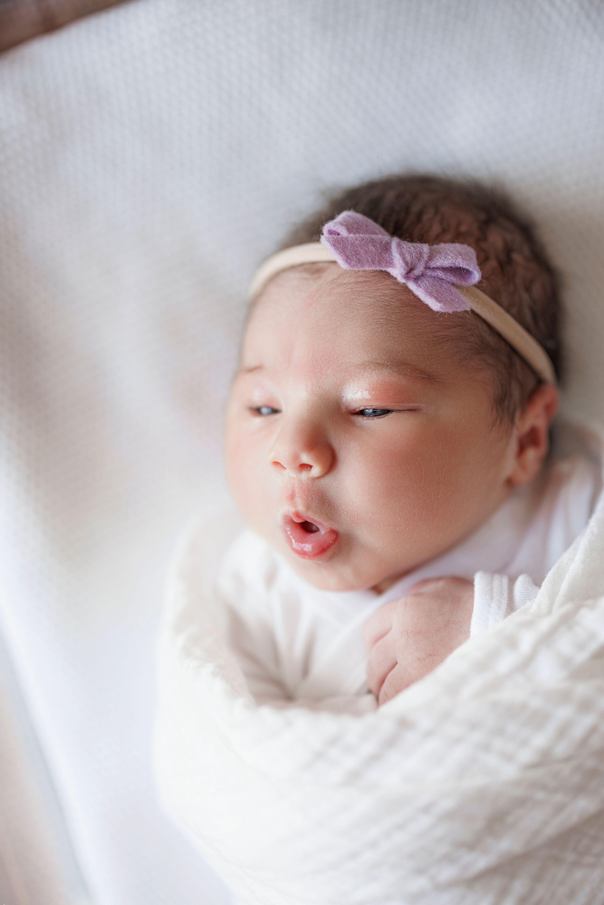Sweet newborn baby swaddled in white blanket with purple bow headband resting peacefully.