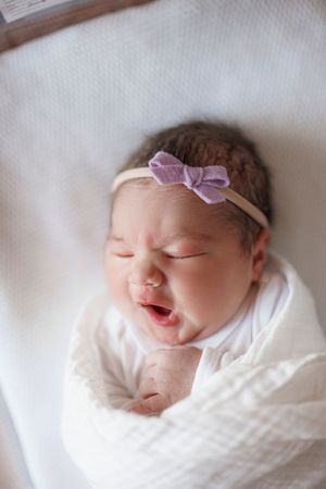 Adorable newborn wearing purple headband and white swaddle making sweet expressions during photoshoot.