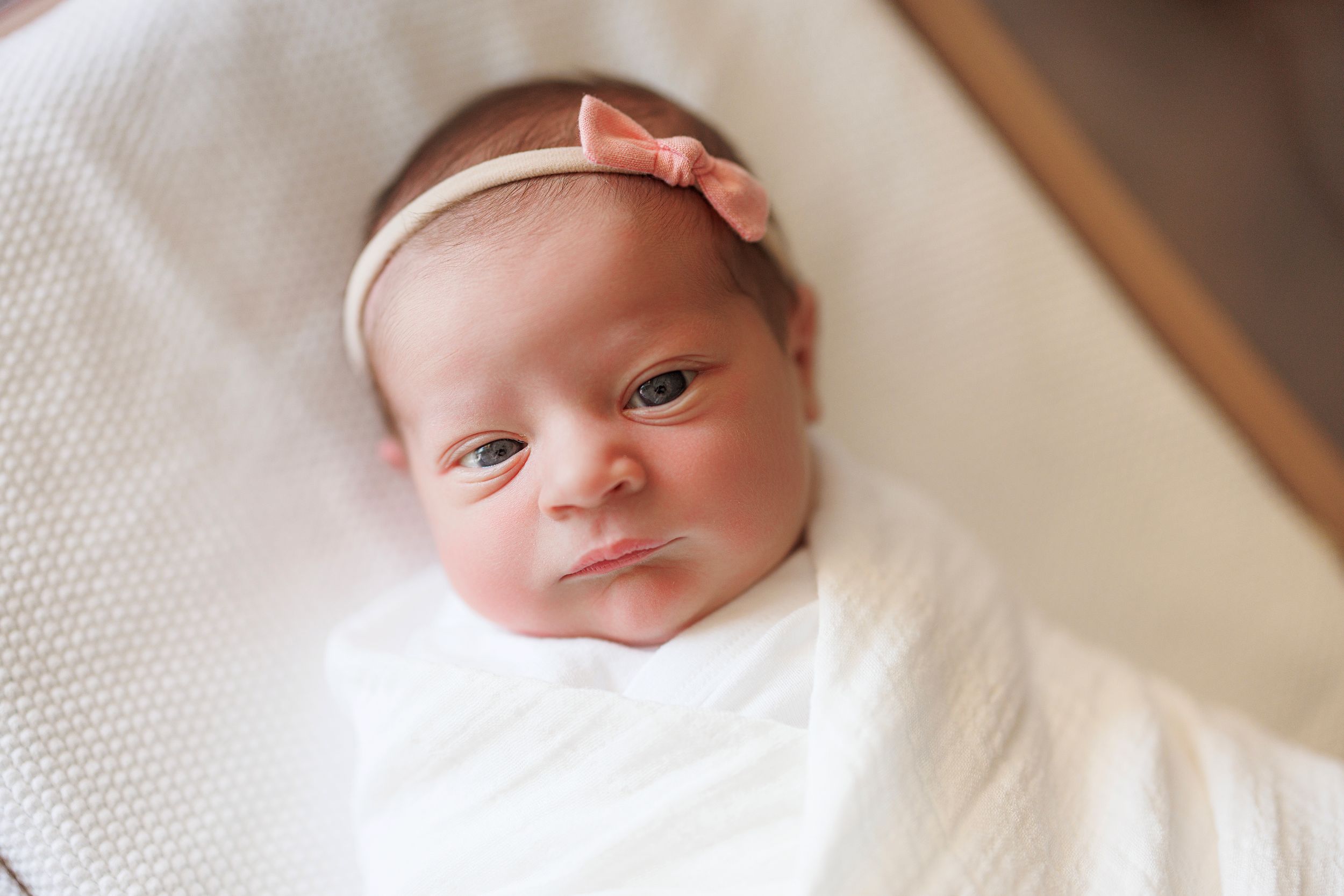 A newborn baby wearing a pink headband lies on white bedding while looking at the camera.