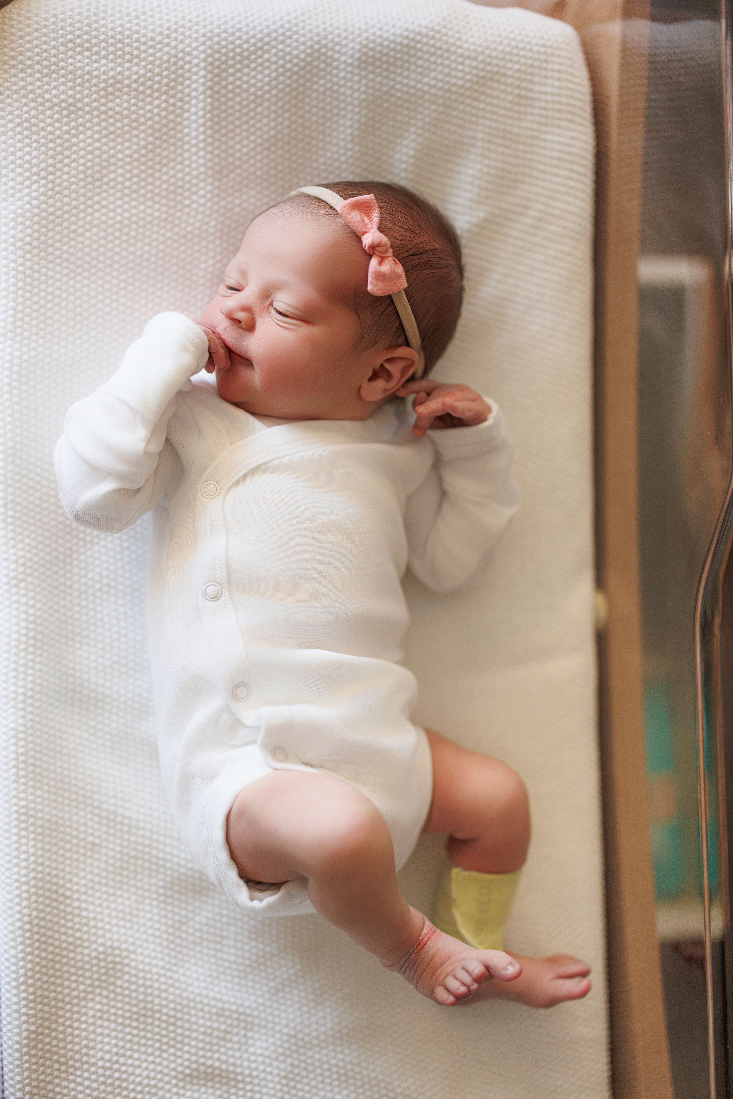Sleeping newborn baby wearing white onesie rests peacefully on soft white textured fabric.