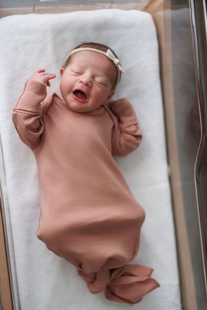 Newborn baby wearing soft peach-colored sleeper lies on white blanket with joyful expression.