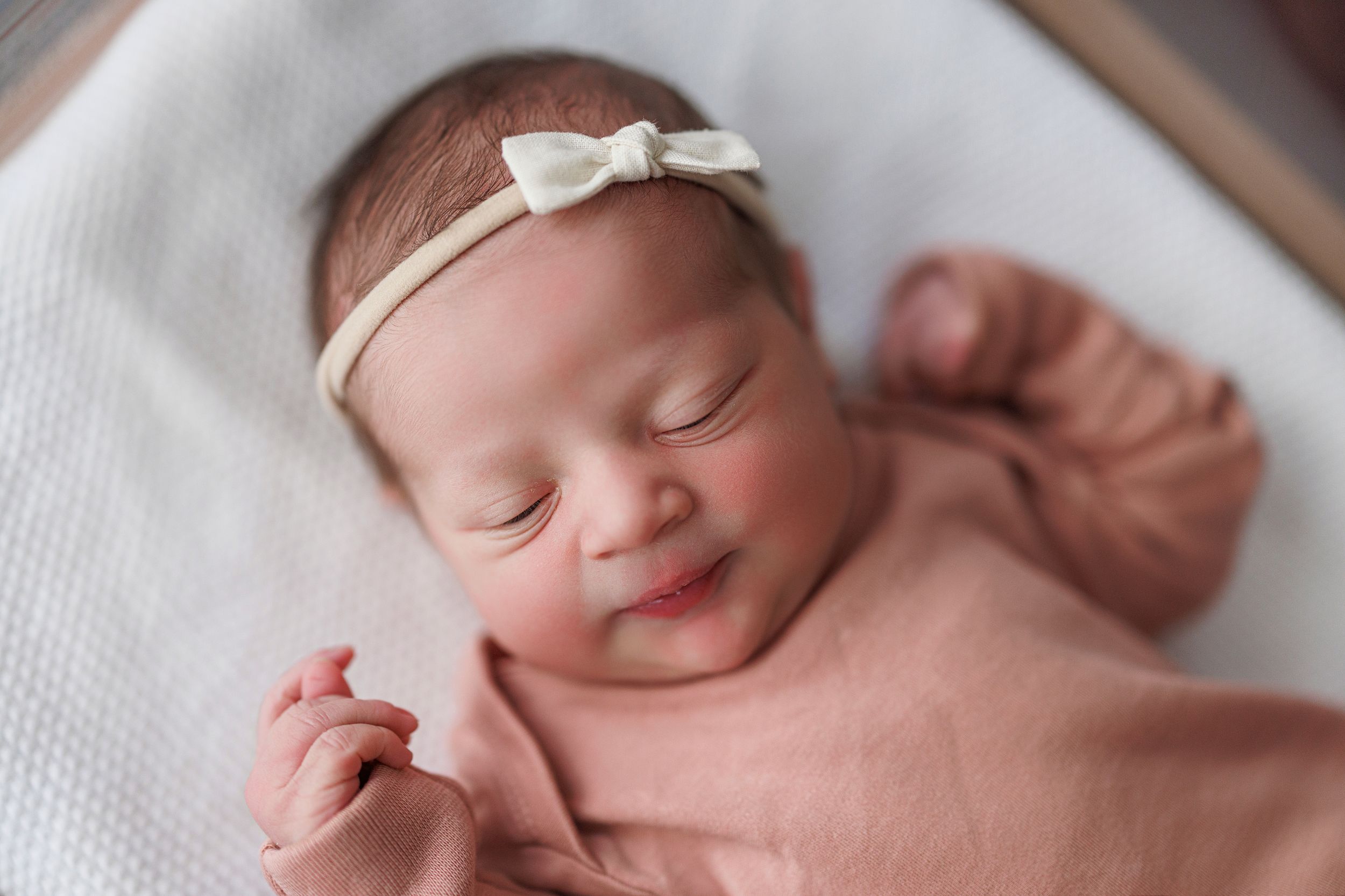 A peaceful sleeping newborn baby wearing a small white bow rests on soft white textured fabric.
