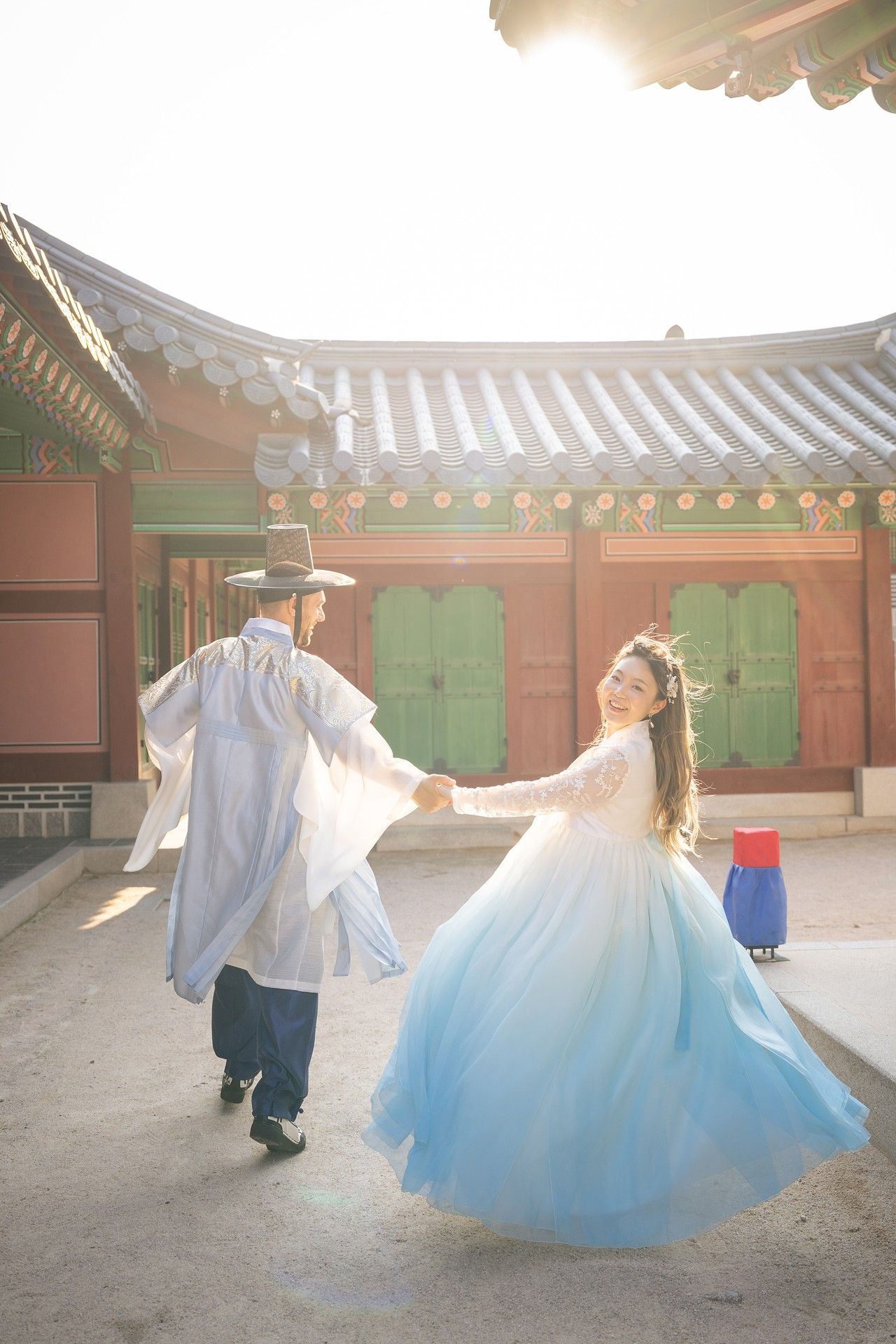 A couple in traditional Korean hanbok attire dances gracefully in a sunlit courtyard of a historic palace.