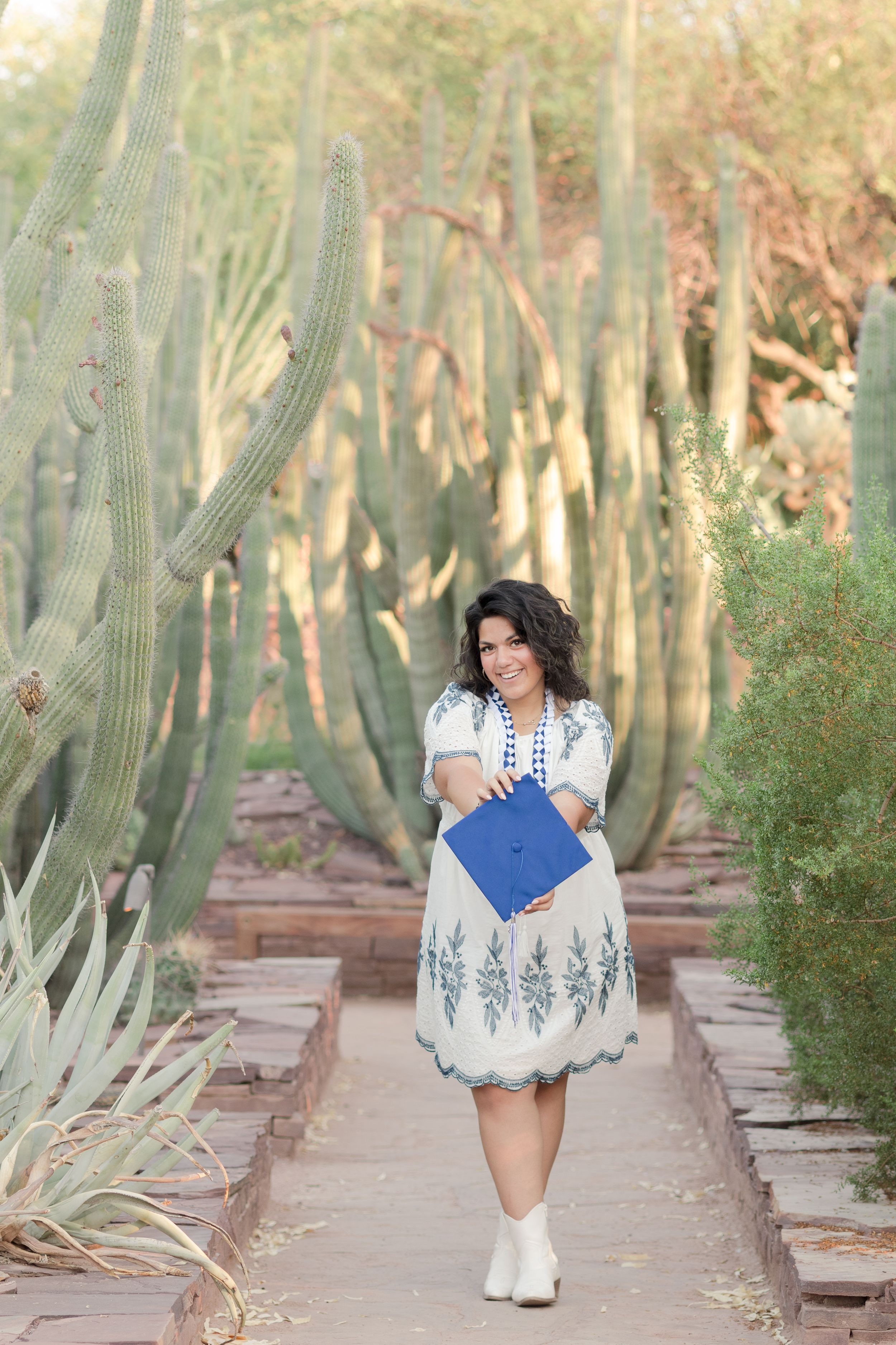 Light and airy senior photo of a girl holding her graduation cap in the middle of cacti at the Desert Botanical Garden in Phoenix, Arizona.