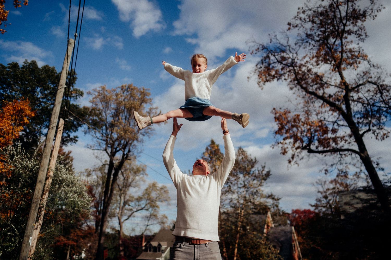 A cheerleader performs a dramatic leap while being supported in an outdoor autumn setting with trees in the background.