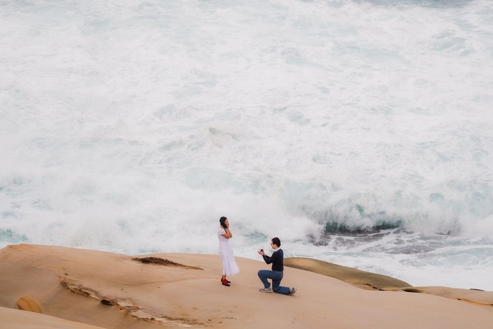A couple proposes on a rocky coastal cliff overlooking dramatic ocean waves crashing against the shore.
