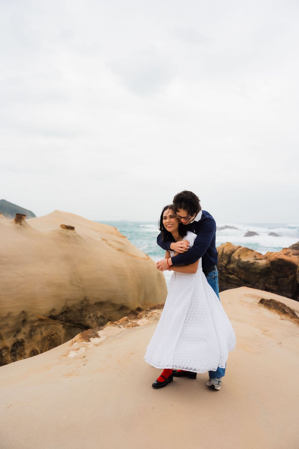 A couple embraces while dancing on sandy coastal rocks with ocean waves and cloudy skies in the background.