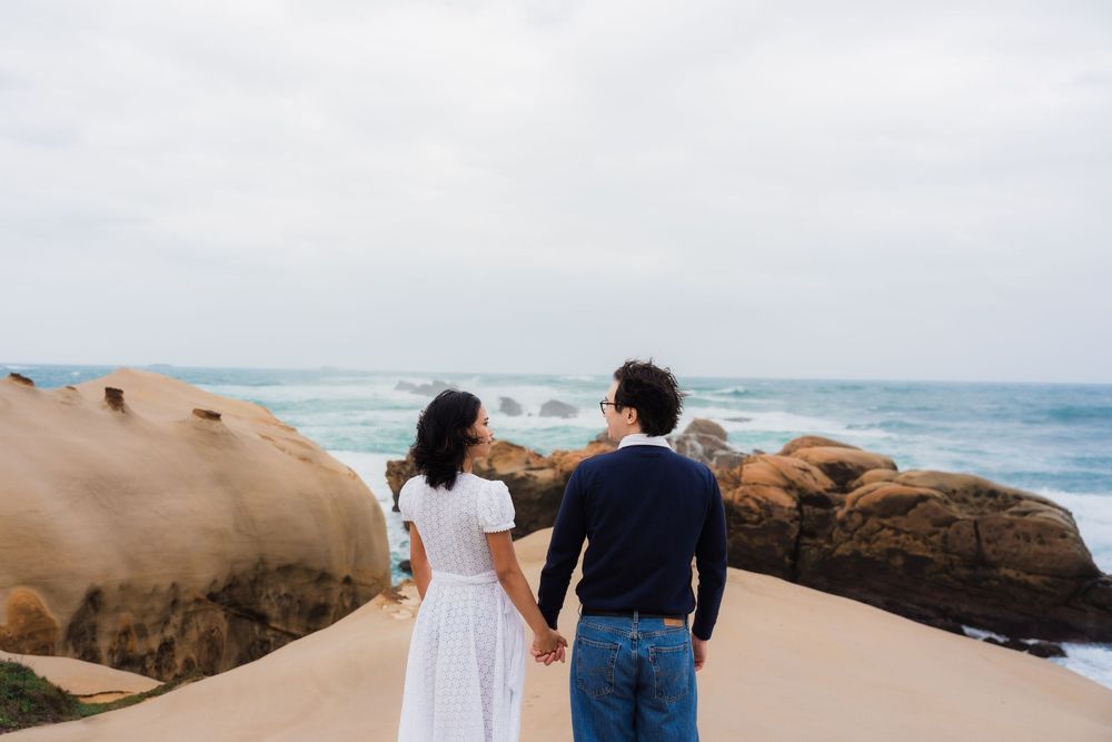 A couple holding hands while walking on a rocky beach with ocean waves in the background.