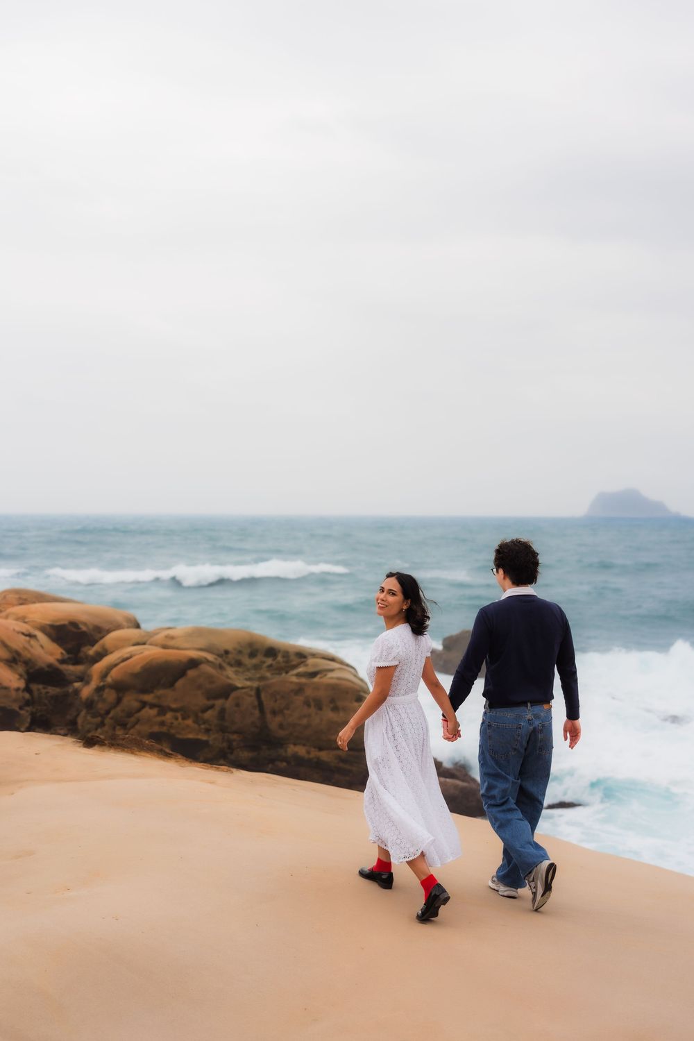 A couple walks along a scenic rocky coastline with crashing waves and misty ocean views in the background.