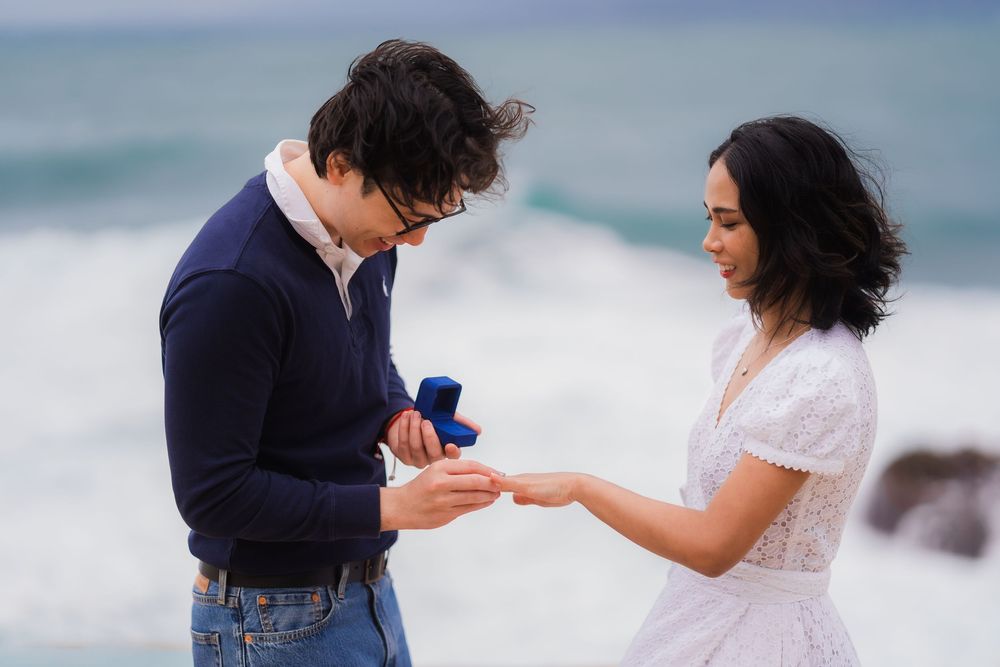 A romantic proposal moment unfolds on a beach with crashing waves in the background.