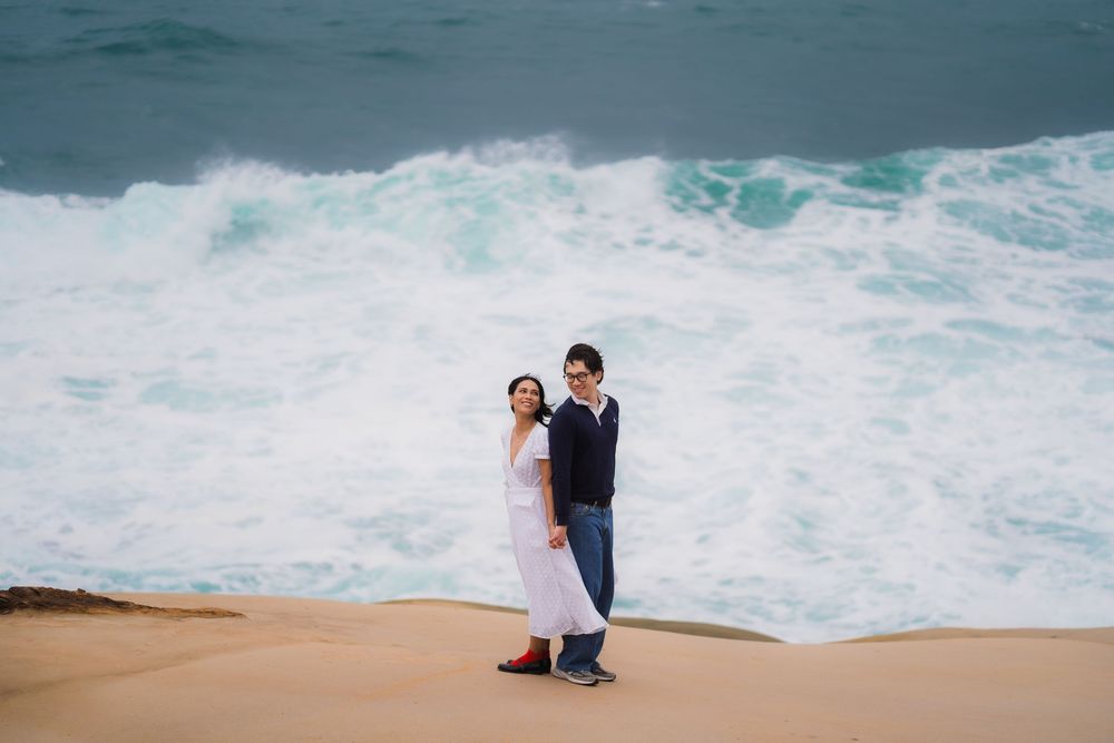 A couple stands on a sandy beach watching powerful ocean waves crash against the shore during sunset.