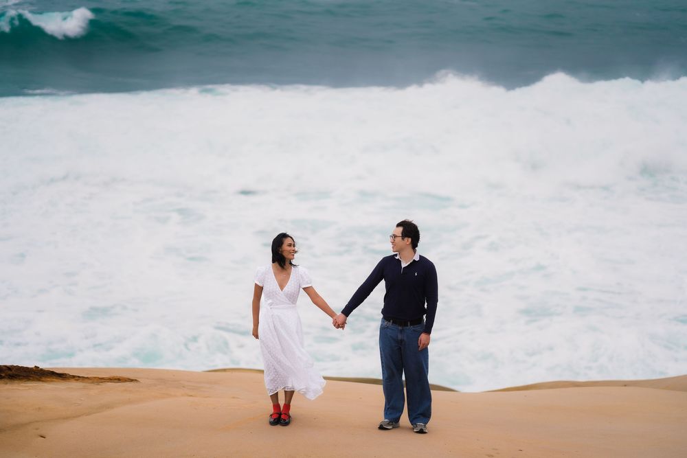 A couple stands hand in hand on a beach with dramatic crashing waves behind them.