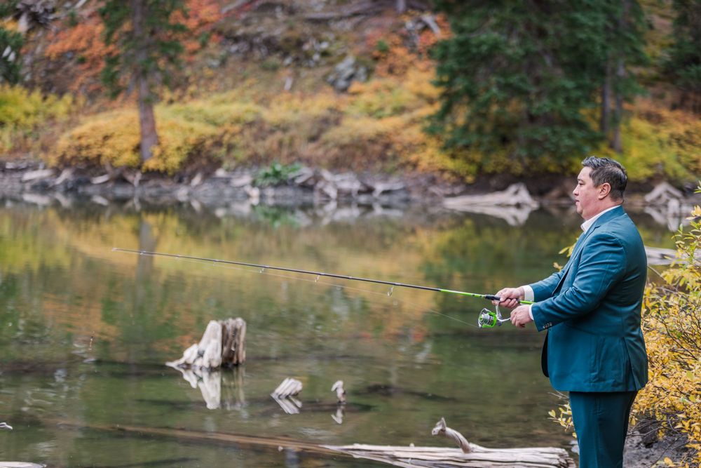 A person in a blue jacket fly fishing at a tranquil lake surrounded by autumn foliage and evergreen trees.