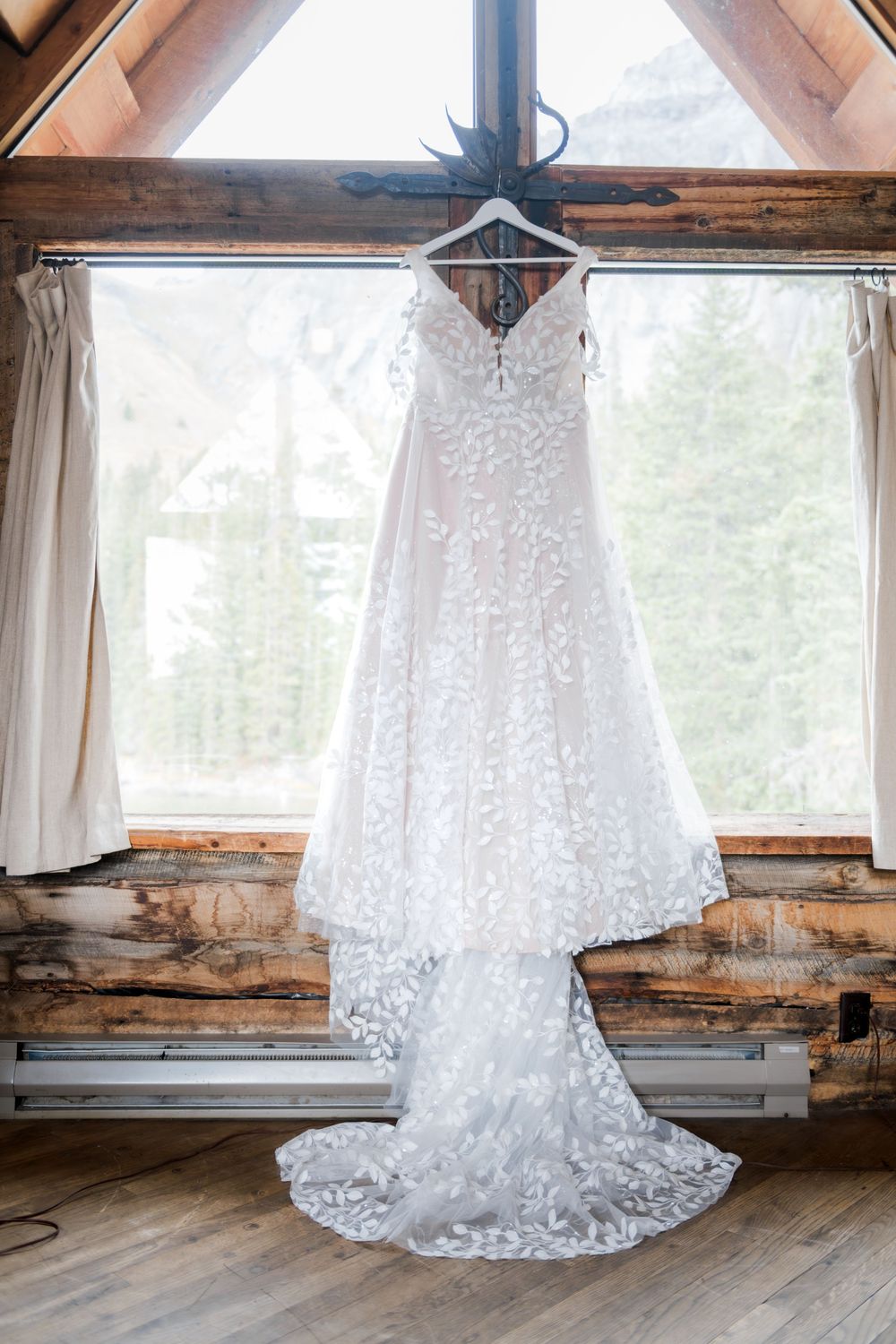 A beautiful white lace wedding dress with a long train hangs from a wooden beam in front of a rustic cabin window.