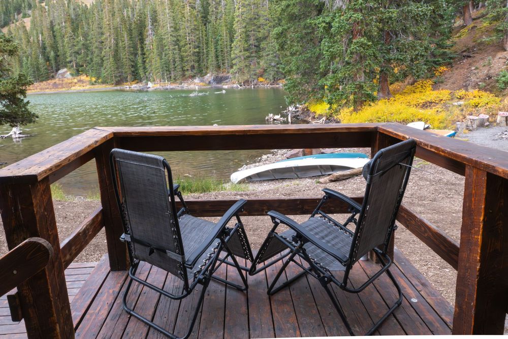 A pair of deck chairs sits on the wooden deck of a rustic cabin overlooking a mountain lake surrounded by pine forest near Telluride.