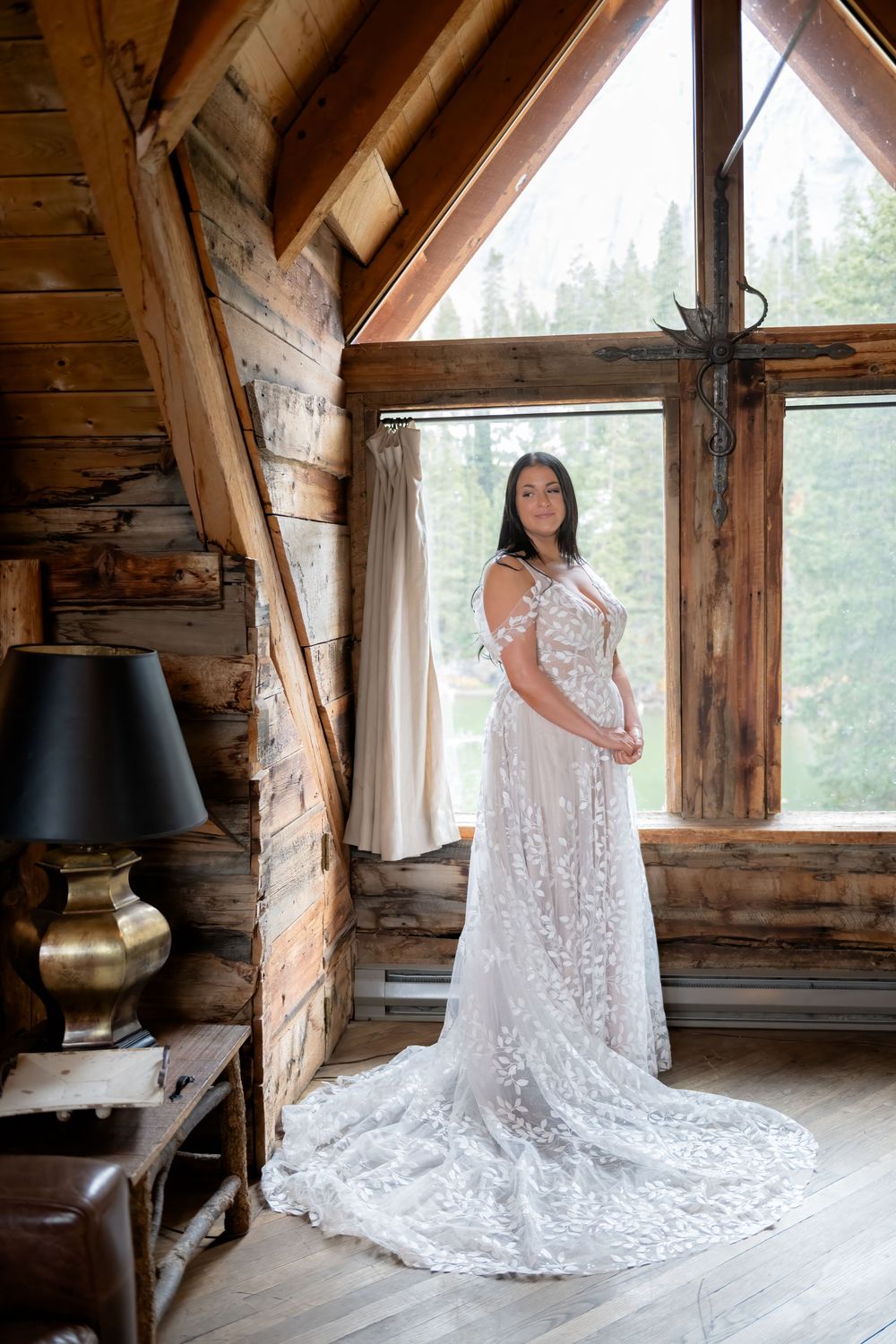 A bride in flowing lace wedding gown with a long train stands by the window in rustic cabin near Telluride with a stone wall backdrop.