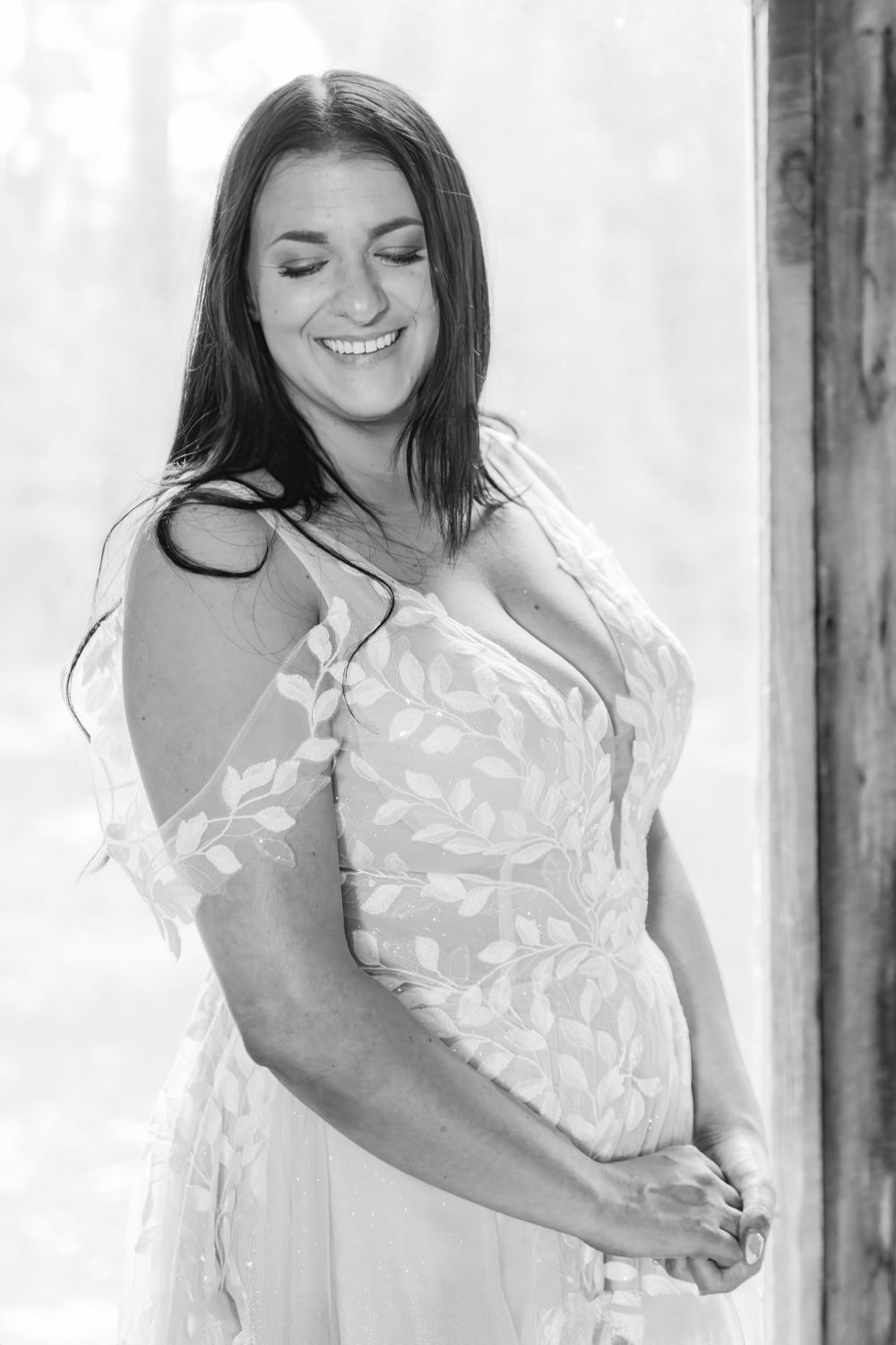 A bride in a white lace wedding dress  smiles brightly in front of a window with natural light. She is looking over her shoulder.