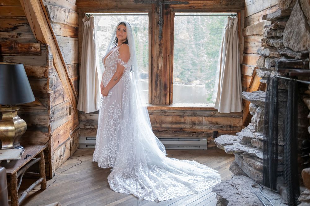 A bride in a flowing lace wedding gown with long train stands by rustic cabin windows in natural light.