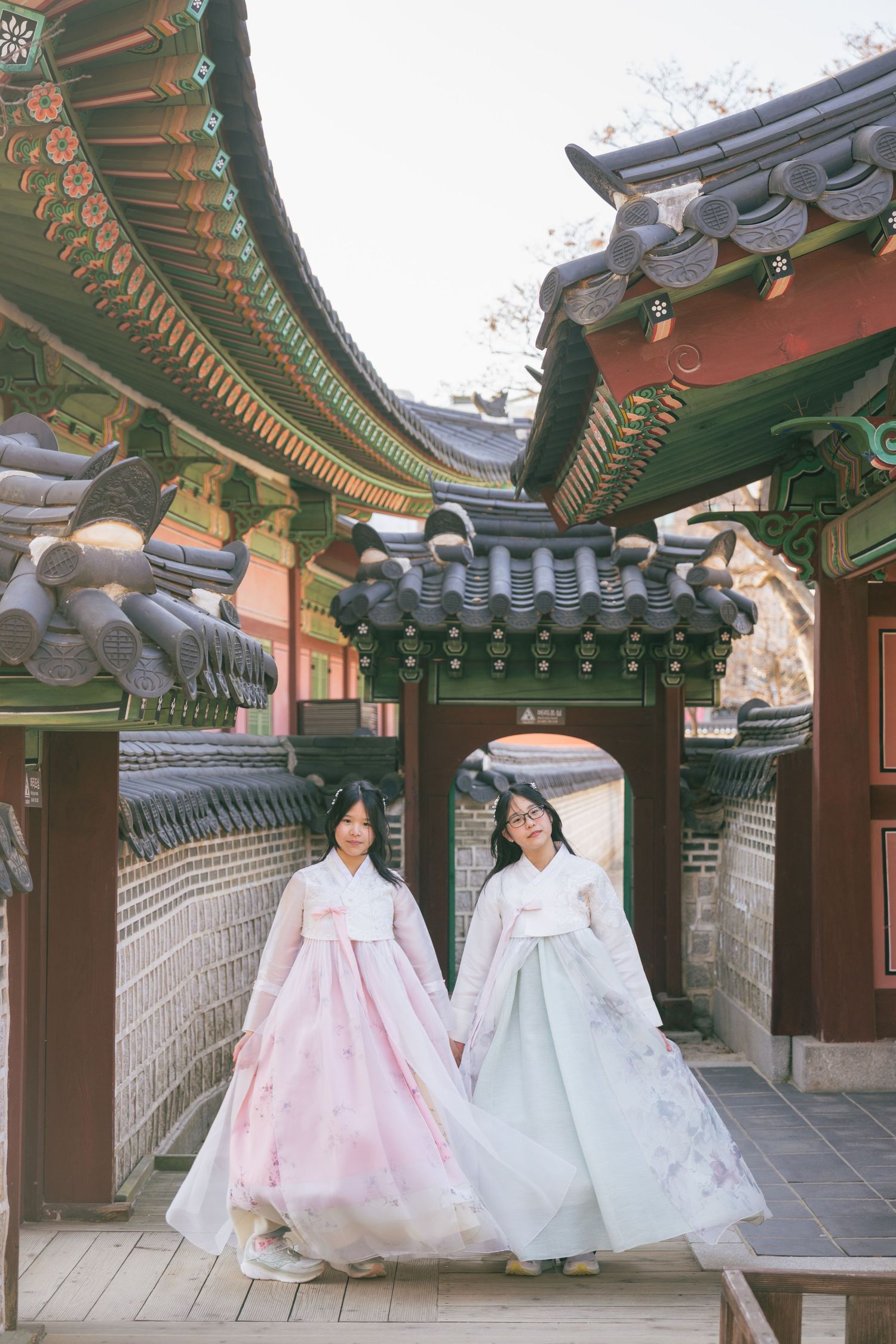 Two people in traditional Korean hanbok dresses walk along an ornate temple corridor with decorative roofs and architecture.