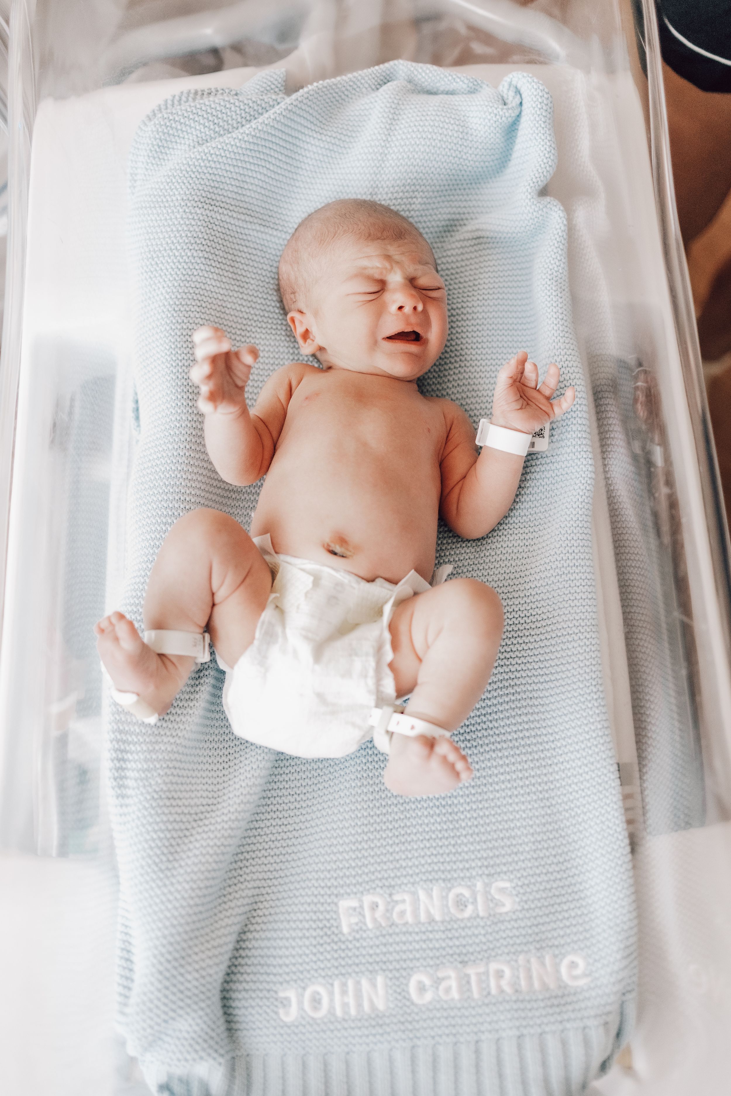 Newborn baby lying on light blue blanket embroidered with 'Francis John Catring' text in hospital bassinet.