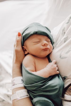 A peaceful sleeping newborn baby wearing a soft green hat rests on white hospital bedding.