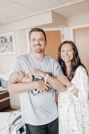 A couple stands together in a bright room holding their newborn baby wrapped in a teal swaddle.