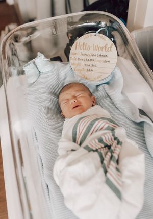A sleeping newborn baby wearing a white onesie rests peacefully on soft gray bedding in a nursery setting.
