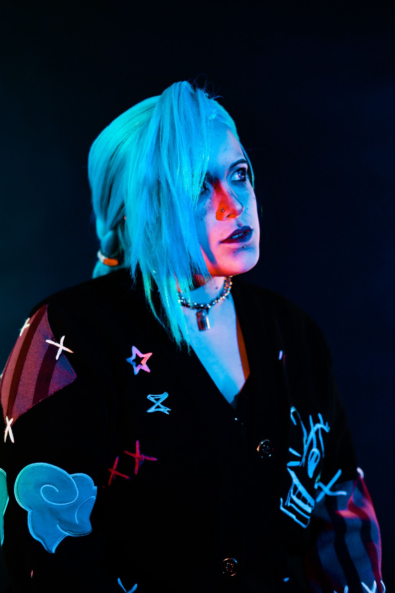 A person with bright blue hair wearing a black jacket with colorful patches poses against a dark background in neon lighting.