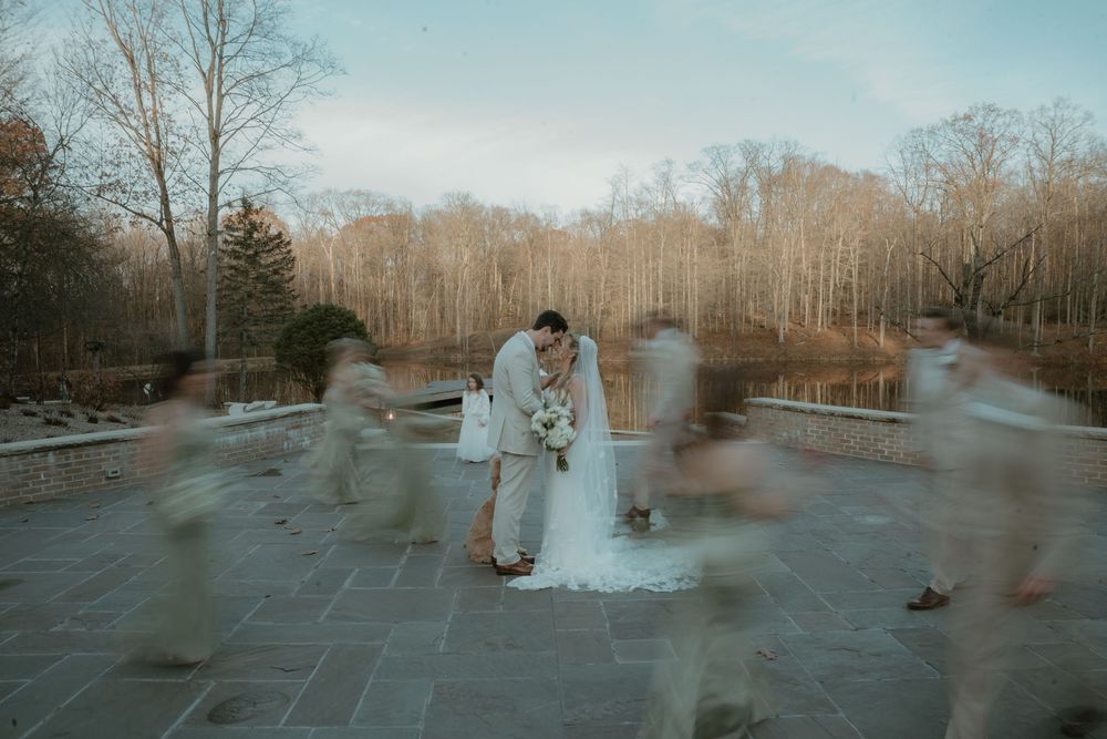 Bride and groom share a kiss while wedding party creates motion blur effect on stone patio surrounded by trees.