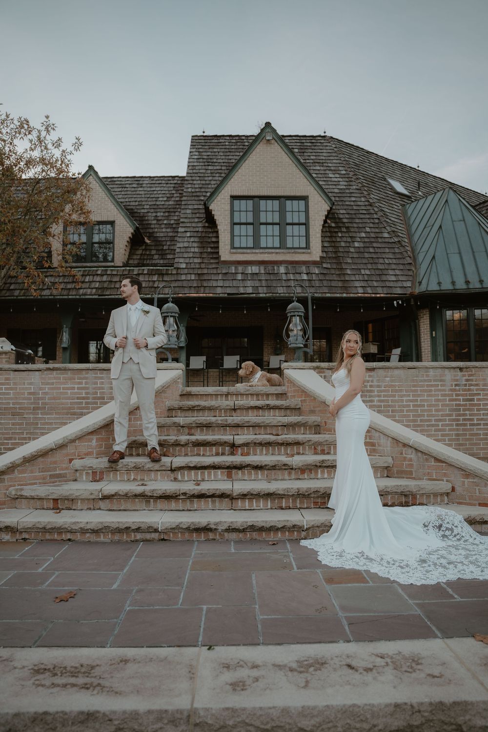Bride in fitted white gown and groom in tan suit pose on brick steps outside elegant Tudor-style wedding venue.