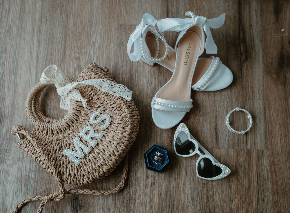 Wedding flat lay featuring woven MRS bag, white heeled sandals, heart sunglasses, and bridal accessories on wood floor.