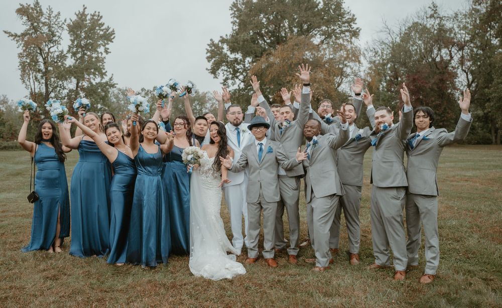 Large wedding party celebrating outdoors, bridesmaids in blue dresses and groomsmen in gray suits cheering together.