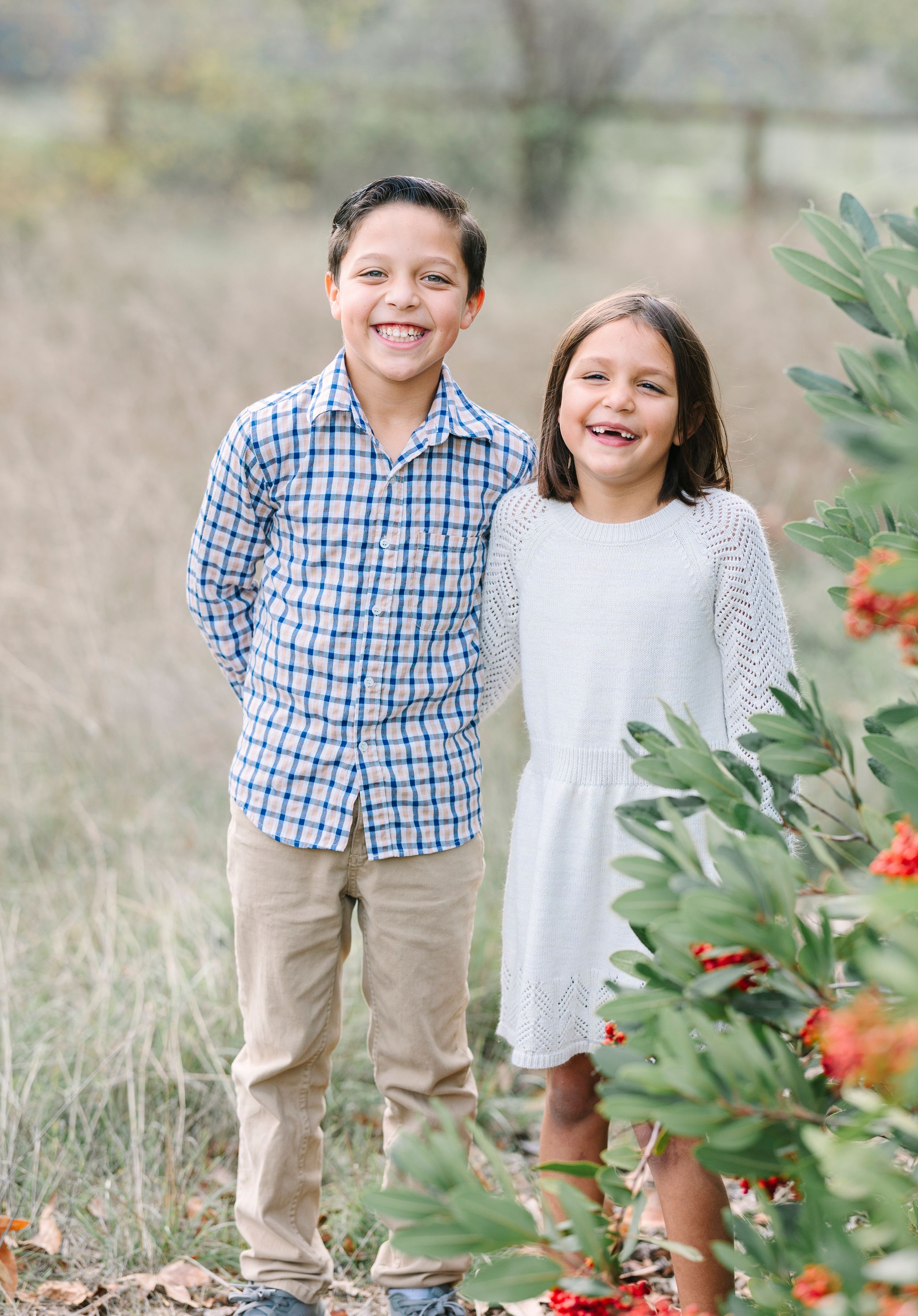 Two children in coordinated outfits standing by red flowering bushes outdoors.