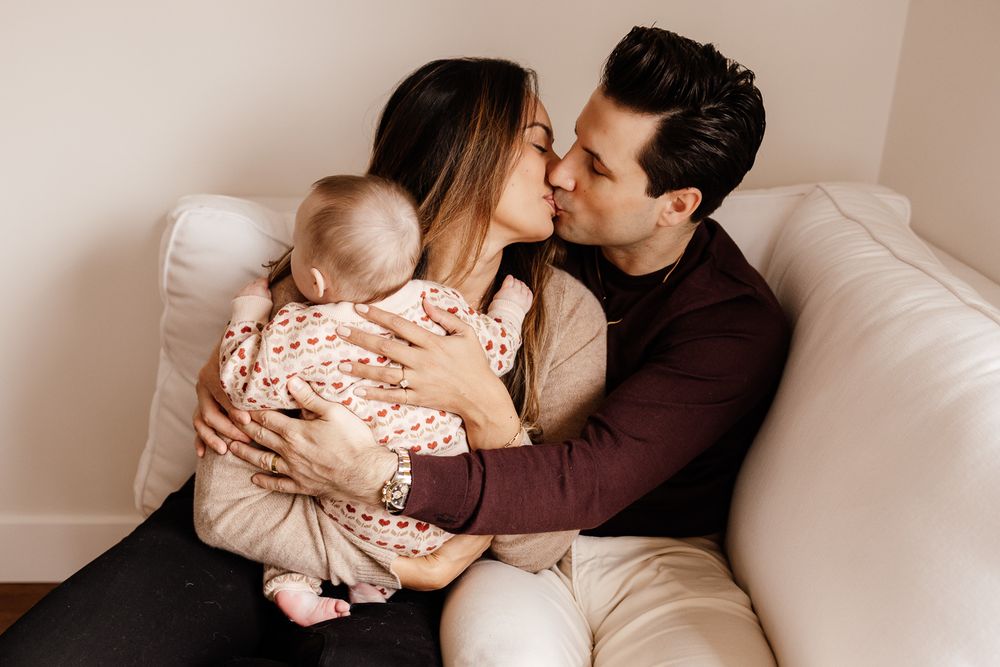 A couple kiss on their white chair while holding their baby